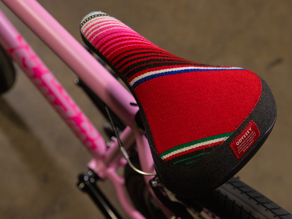 Close-up of the Sunday High C 29 Inch Bike—a pink cruiser bicycle with a vibrant, striped red seat featuring a small Odyssey brand label—set against a blurry brown background. This bike highlights its sturdy chromoly frame, offering both style and durability.