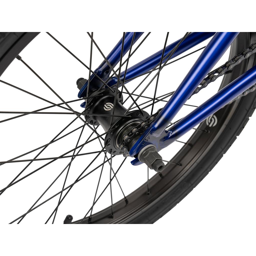 Close-up of the Wethepeople CRS FC 20 Inch Bike’s rear wheel, showing black spokes, chain, hub, and a metallic blue entry-level frame with a freecoaster hub.
