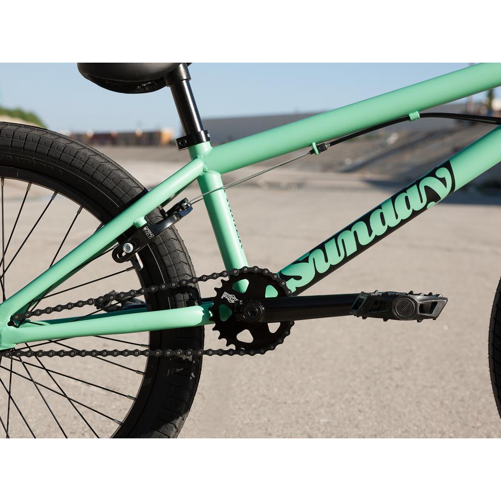 Close-up of the Sunday Primer Park 20 Inch Bike frame highlights its chain, pedals, crankset, and "Sunday" logo, with a blurred skatepark backdrop—showcasing key details of this park BMX bike.