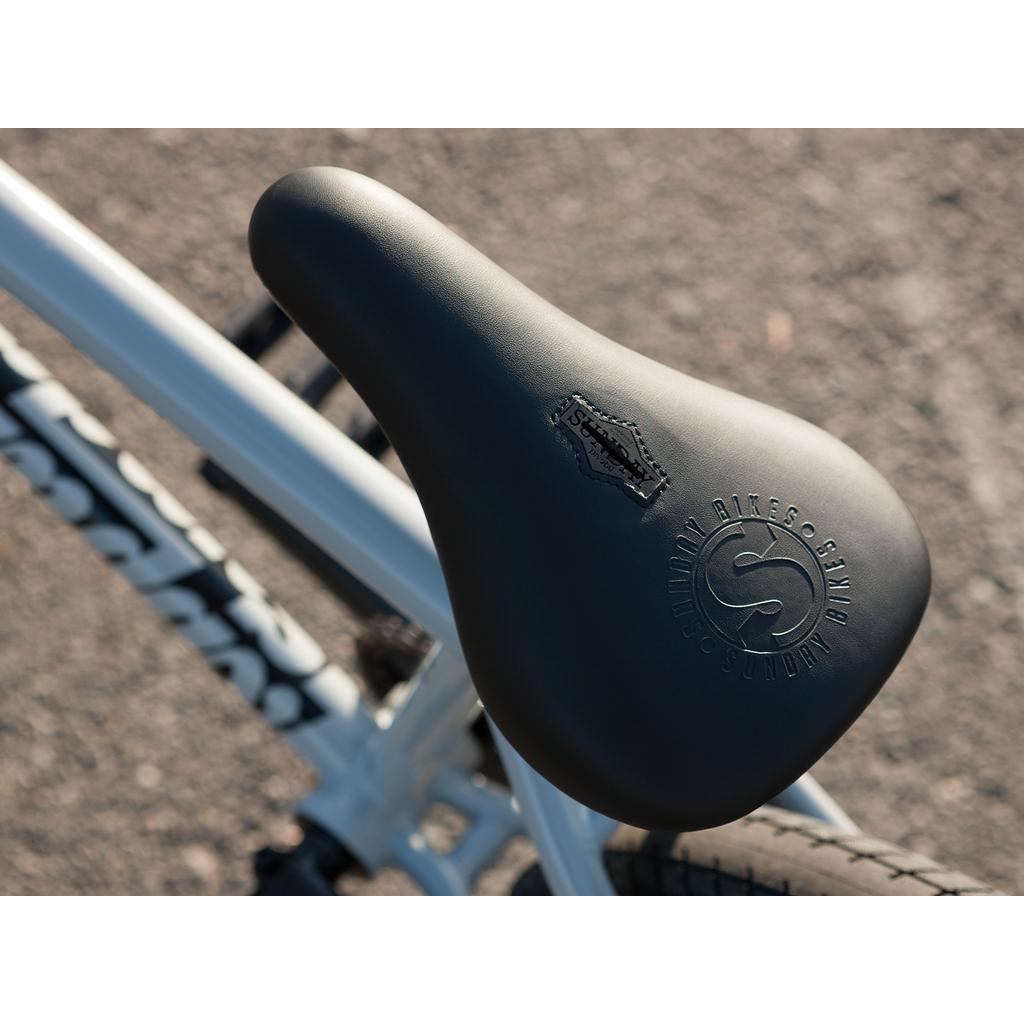 Close-up of the black seat with embossed branding on the Sunday EX Erik Elstran 20 Inch Bike, featuring a white 100% chromoly frame, photographed outdoors on pavement.