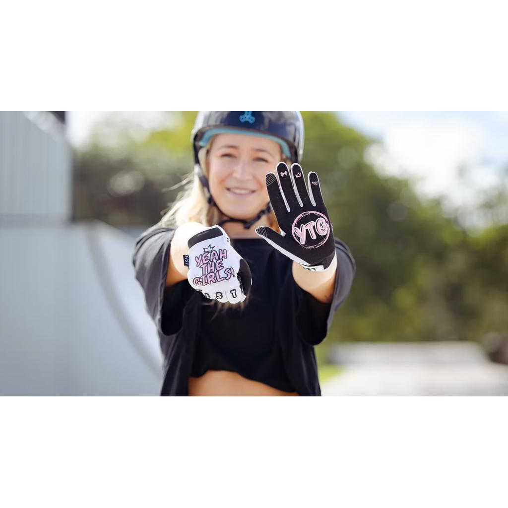Wearing a helmet, a person smiles and displays the YTG x Fist Adult Gloves—one palm says "YEAH THE GIRLS," the other "YTG"—at an outdoor skate park, celebrating female freestyle BMX riders and culture.