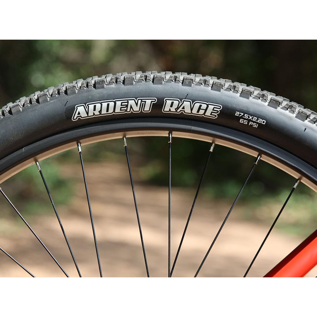 Close-up of an "ARDENT RACE" bicycle tire, sidewall marked 27.5x2.20 and 65 PSI—perfect for the Fairdale Taj 27.5 Inch Bike (2023)—set against a blurred outdoor background.