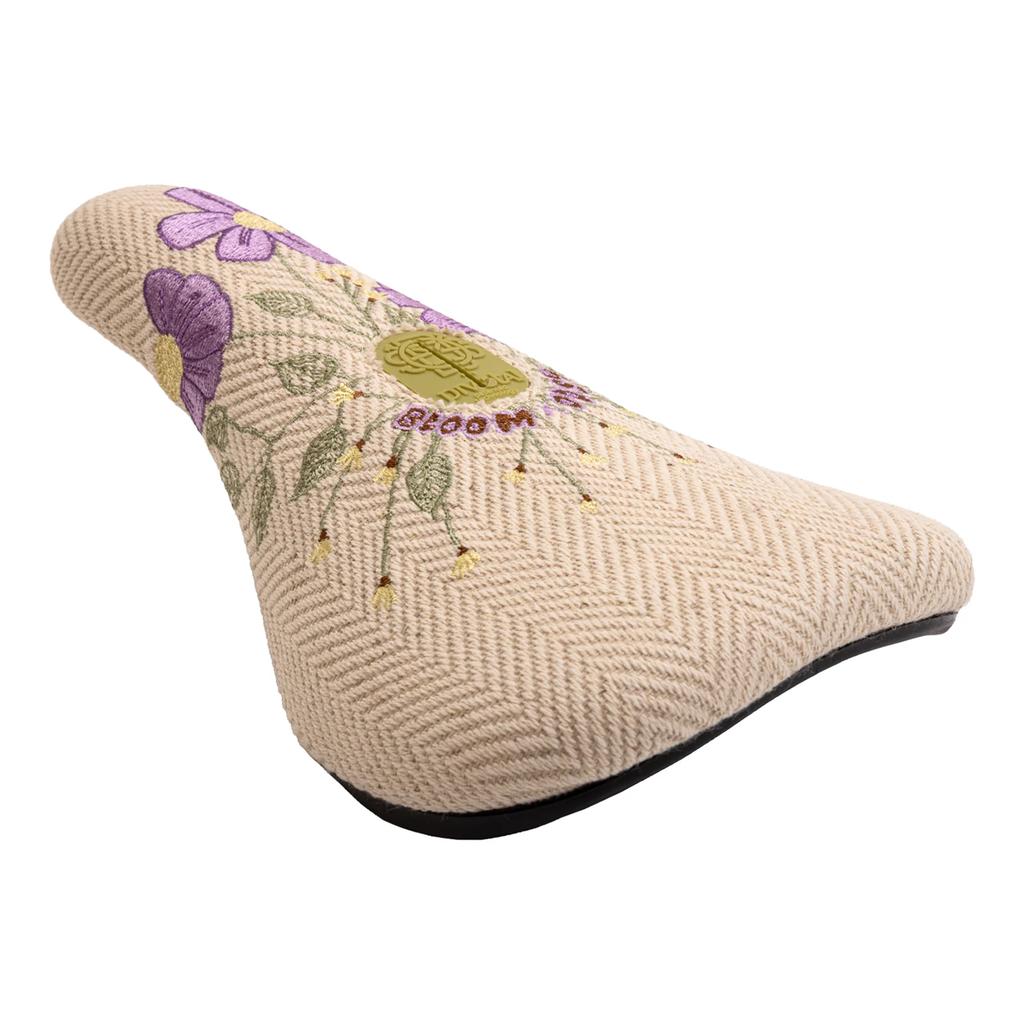 The Odyssey Bloom V2 Pivotal Seat features beige herringbone fabric, embroidered purple flowers, and "bloom" and "ride" text—designed to inspire women riders.