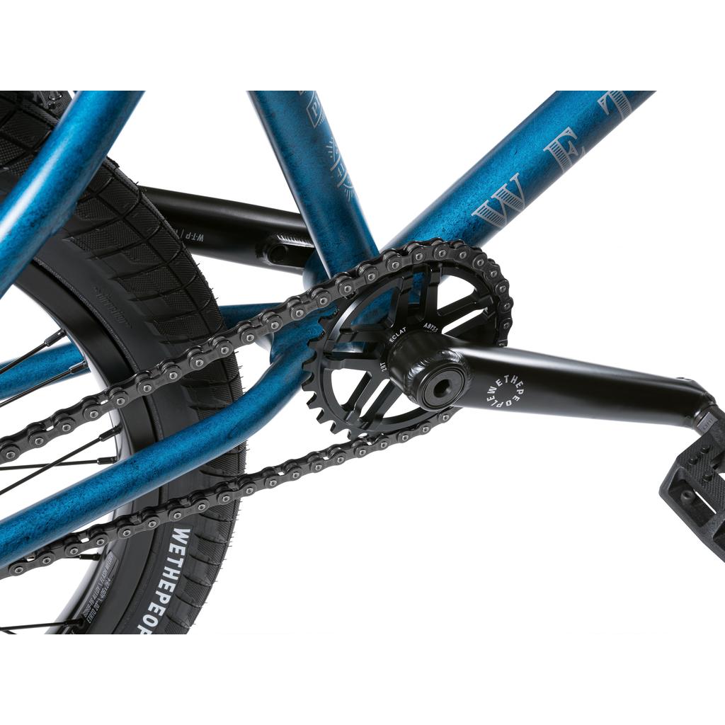Close-up of a BMX drivetrain showing the chain, chainring, crank arms, rear tire, and blue frame with "WETHEPEOPLE" branding from the Wethepeople Revolver 20 Inch Bike.