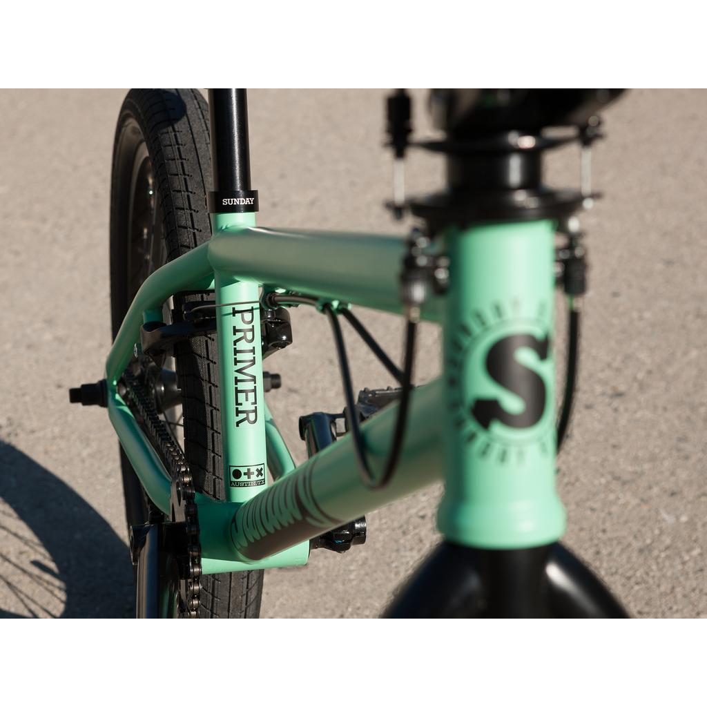 Close-up of the mint green Sunday Primer Park 20 Inch Bike frame, labeled "PRIMER" and "SUNDAY," highlighting the rear tire, part of the front fork, and handlebar stem on this BMX park bike.