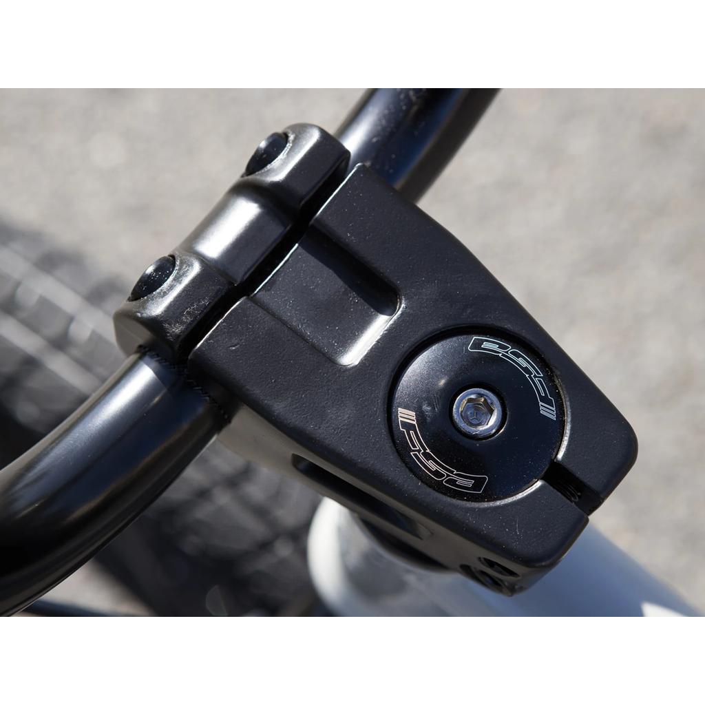 Close-up of a Sunday Blueprint 20 Inch Bike (2023) showing its black stem and handlebars with a visible bolt and logo, photographed outdoors in daylight—compatible with Odyssey Springfield brakes for added control.