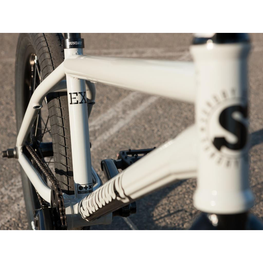 Close-up of a white Sunday EX Erik Elstran 20 Inch Bike with a 100% chromoly frame, highlighting the rear wheel, pedal, and "S" logo on the head tube, parked on an asphalt surface.