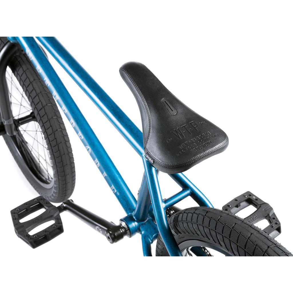 Close-up of a blue Wethepeople Revolver 20 Inch Bike BMX, highlighting the black seat, rear tire, pedals, and part of the bike frame on a white background.