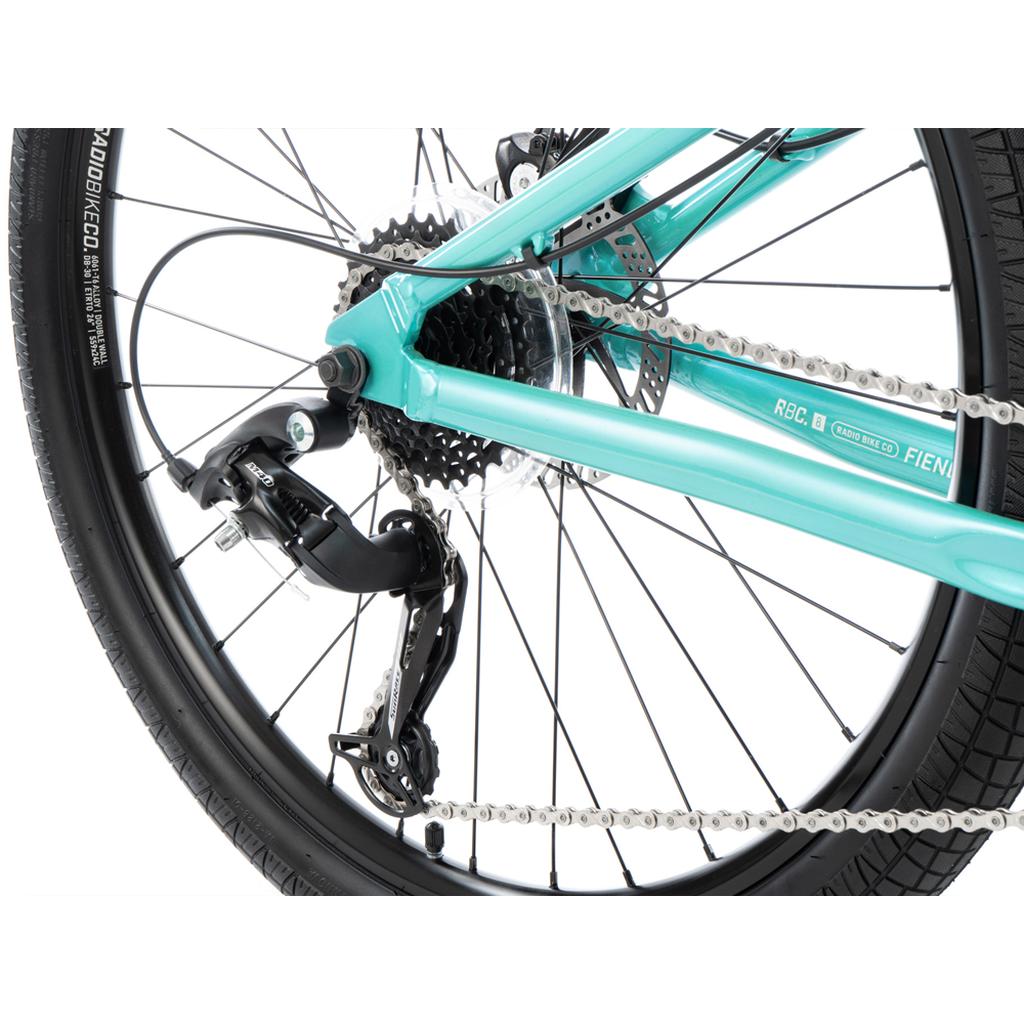 Close-up of the Radio Fiend 26 Inch Bike's rear wheel, showcasing the derailleur, chain, cassette, and a section of its turquoise alloy BMX frame.