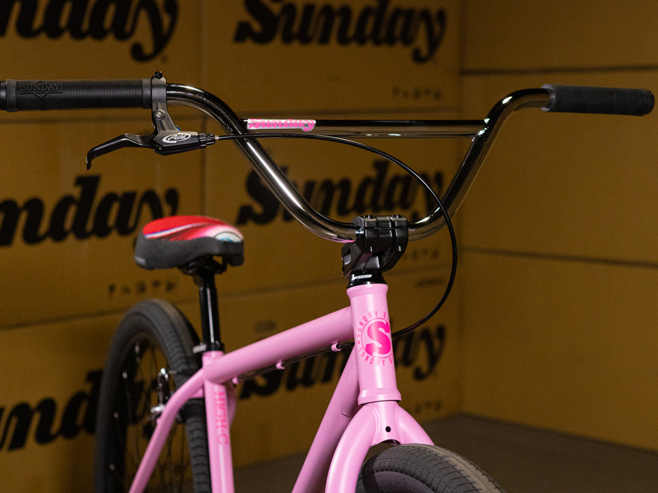 Close-up of the Sunday High C 29 Inch Bike in pink, showcasing its black handlebars and black-and-red seat, set against a backdrop of repeating “Sunday” logos. This stylish cruiser features a robust chromoly frame, delivering both standout looks and exceptional performance.