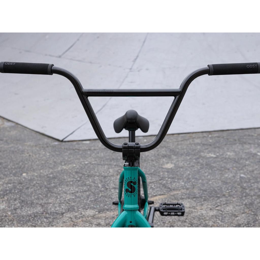 Close-up of the handlebars, seat, and front frame of the Sunday EX Erik Elstran 20 Inch Bike with a teal 100% chromoly frame and Odyssey/Sunday parts, shown on concrete near a skate ramp.