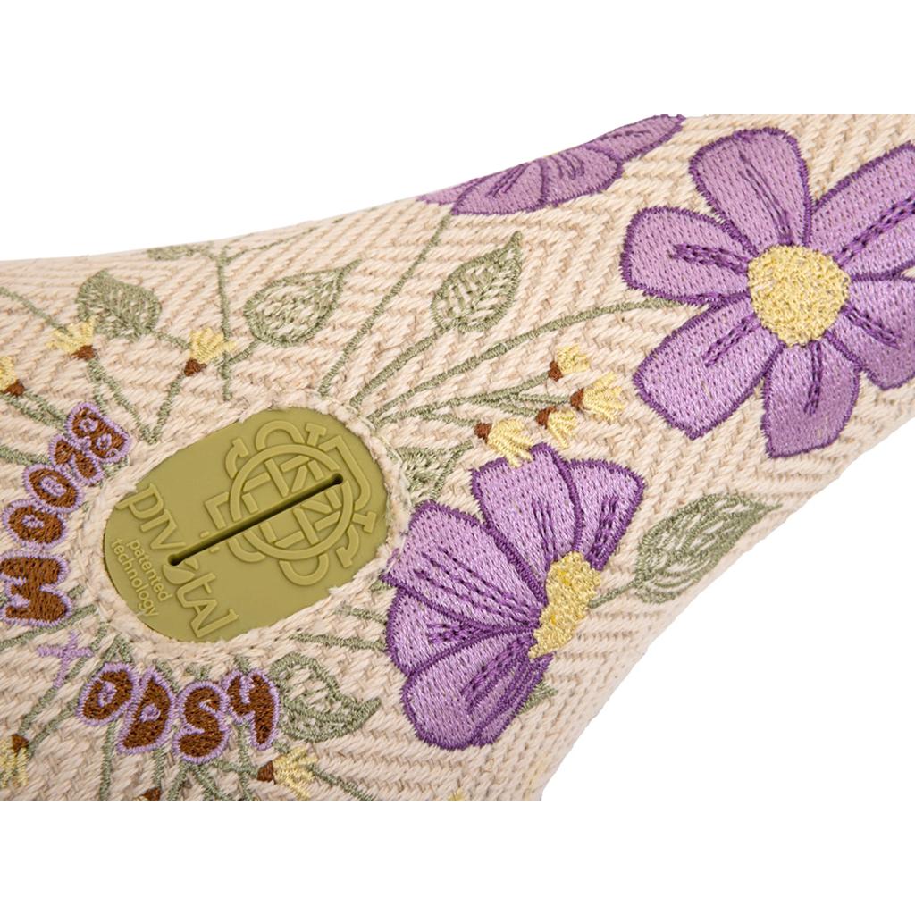 A close-up of the Odyssey Bloom V2 Pivotal Seat shows textured fabric with purple and yellow embroidered flowers and a beige plastic disk labeled "PIN KAI"—ideal for women riders who love detailed seat designs.