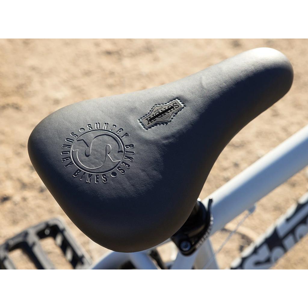 Close-up of a black saddle with textured branding on the Sunday Model C 24 Inch Bike’s white Chromoly frame, pictured outdoors on dirt.