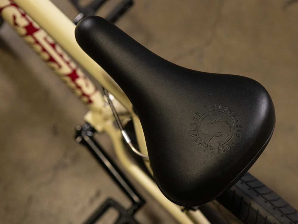 Close-up of a black seat featuring embossed branding, mounted on the cream-colored chromoly frame of the Sunday High C 29 Inch Bike, resting on a concrete floor.