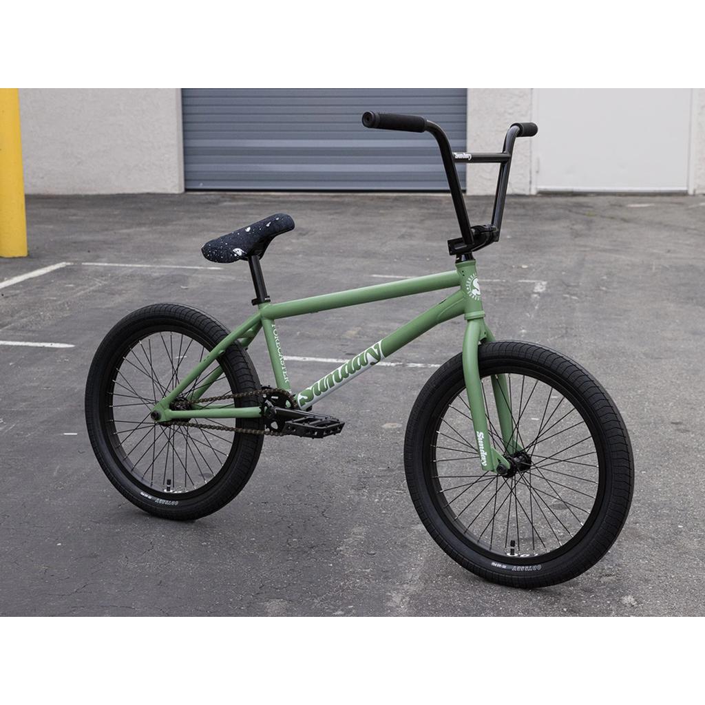 A matte green Sunday Forecaster 20 Inch Bike (2025) / Brett Silva pro-level BMX, featuring black handlebars, seat, and tires, is parked on an empty paved lot near a building with a closed garage door. The 100% chromoly frame adds strength and style.