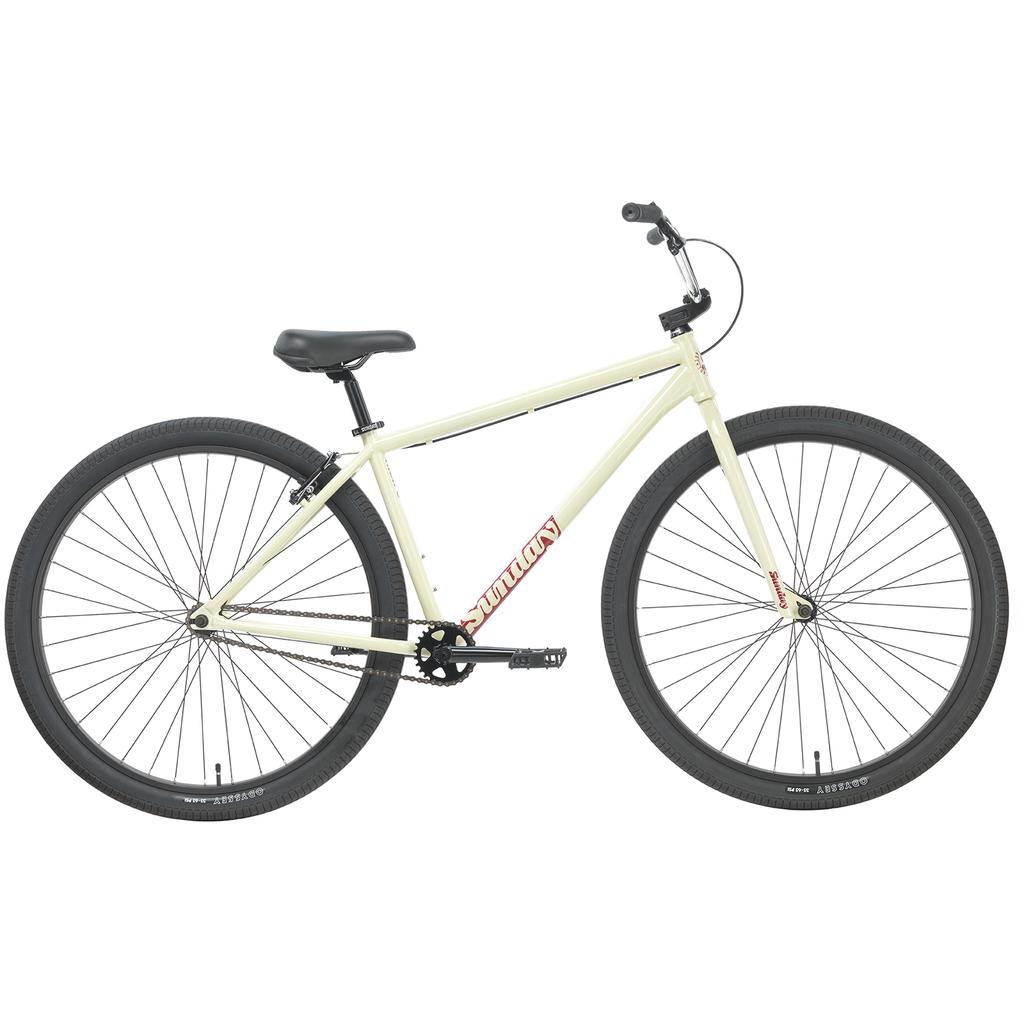 A cream-colored Sunday High C 29 Inch Bike—a single-speed chromoly cruiser with large black tires, flat handlebars, and a black saddle—displayed in profile view on a white background. This model features quality Odyssey components throughout.