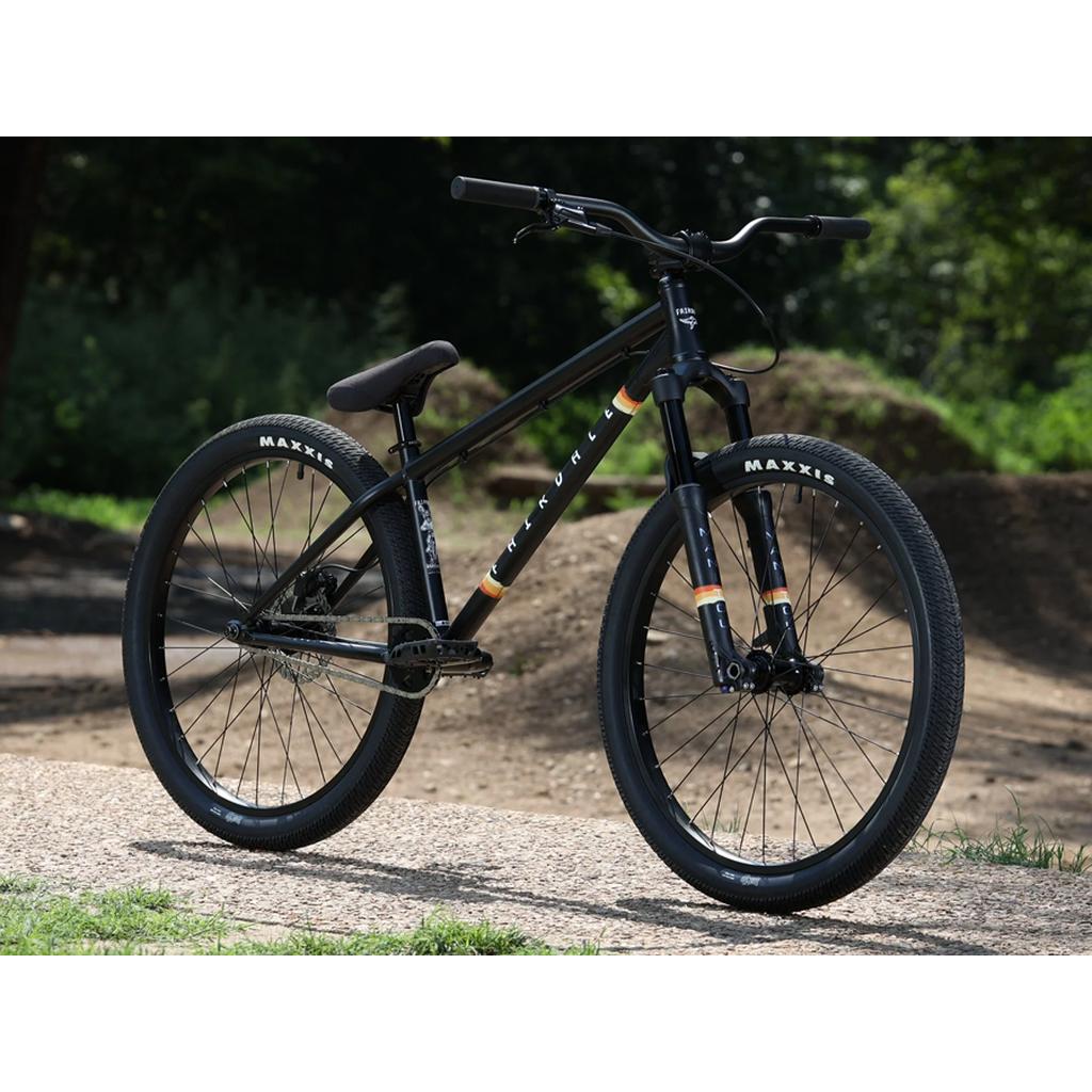 A Fairdale Hareraiser FX 26 Inch Bike with a black DJ frame and Maxxis tires is parked on a concrete path in front of a dirt track and greenery.