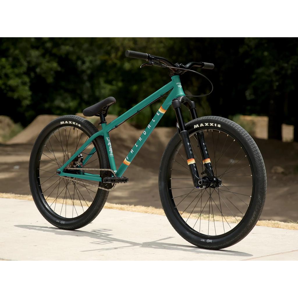 A Fairdale Hareraiser FX 26 Inch Bike in teal, featuring a hardtail DJ frame, black tires, and handlebars, is parked on concrete near a dirt trail and trees.