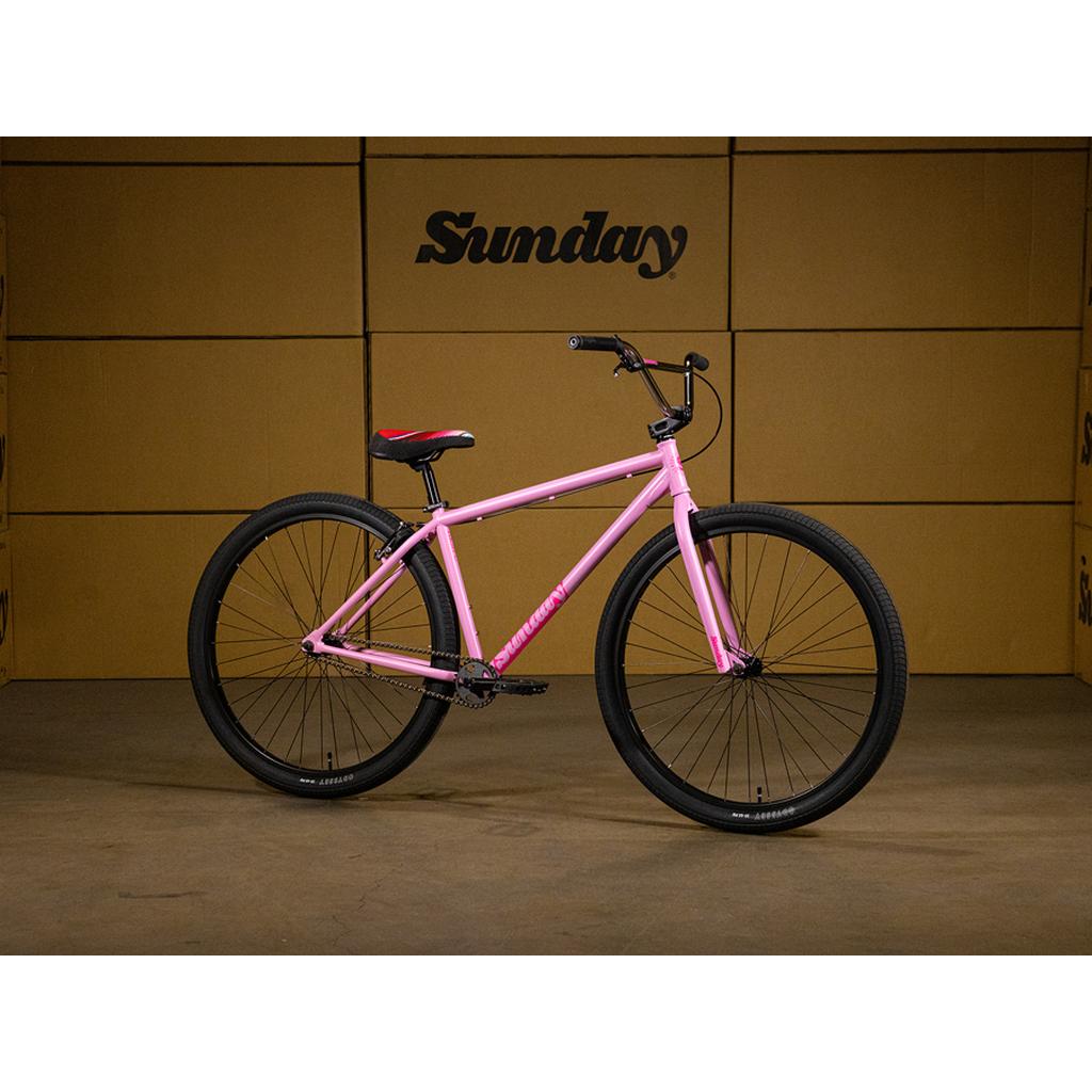 A pink Sunday High C 29 Inch Bike with black tires and a seat featuring red accents is showcased indoors in front of a wall of "Sunday" branded cardboard boxes. This striking display highlights the Sunday High C 29 Inch Bike, recognized for its robust chromoly cruiser frame and high-quality Odyssey components.