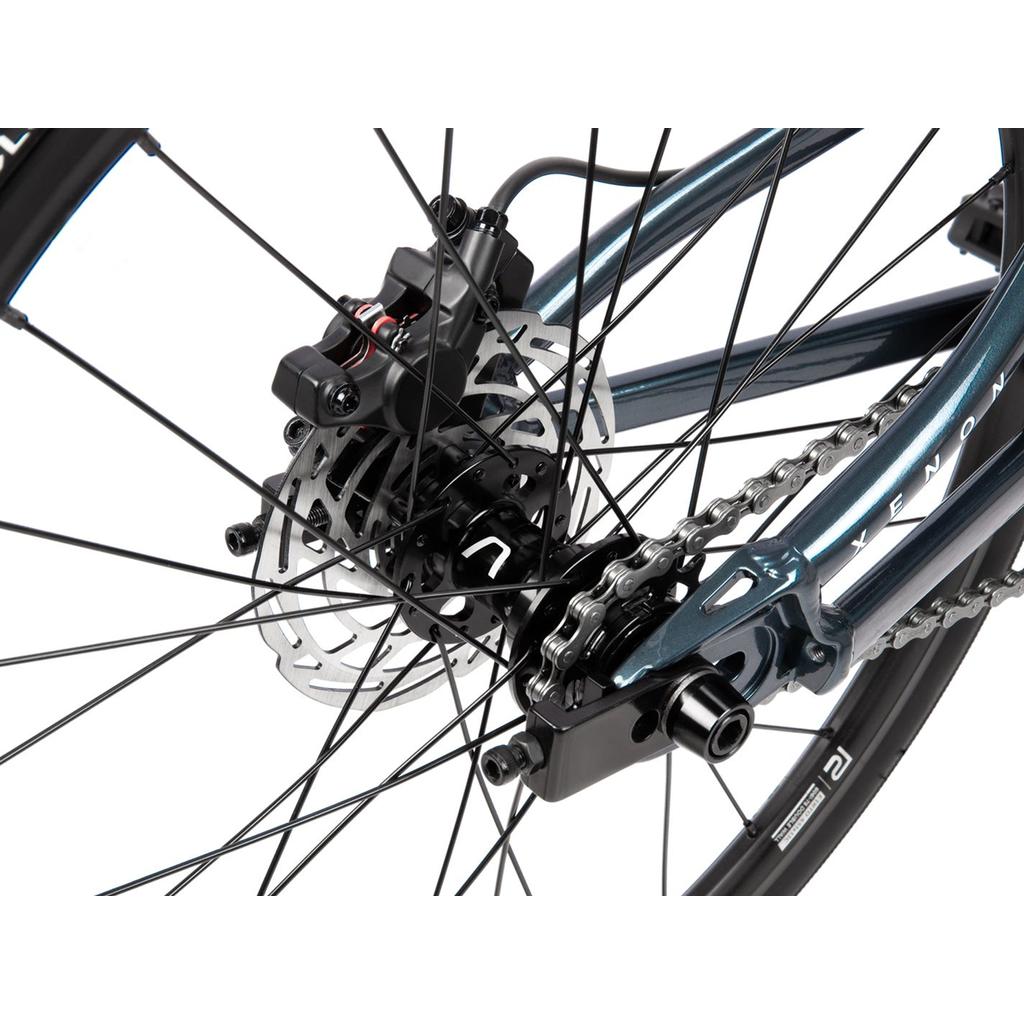 Close-up of the Radio Xenon Junior Disc 20 Inch Bike's rear wheel, highlighting the disc brake, chain, derailleur, spokes, and part of its hydroformed 6061-T6 alloy frame. Partially visible Tioga Power Block tyres underscore the bike’s performance and durability.