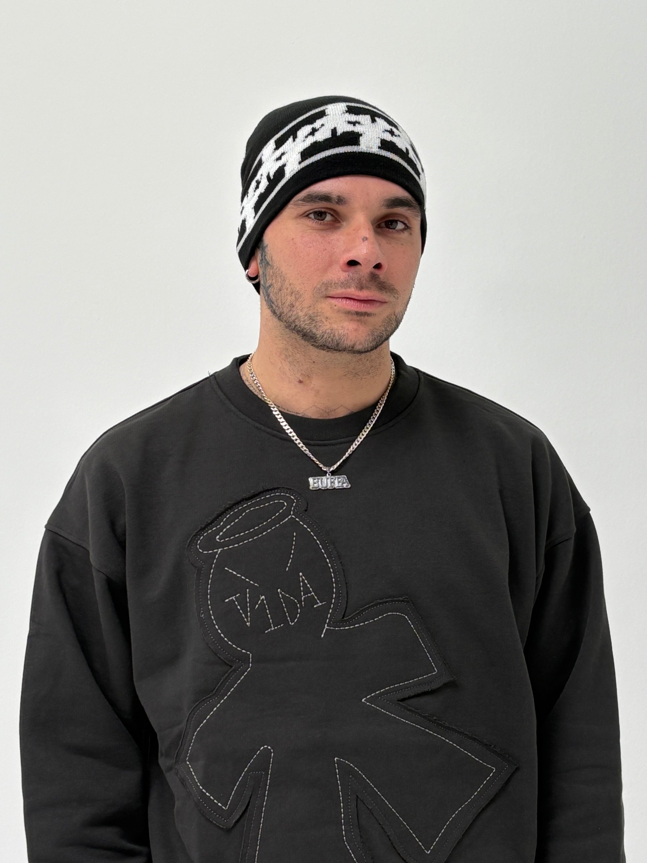 Wearing the V1da Pattern Beanie in black and white low fit, a person pairs it with a black sweatshirt featuring a stitched figure design and a silver necklace, standing against a plain white background.