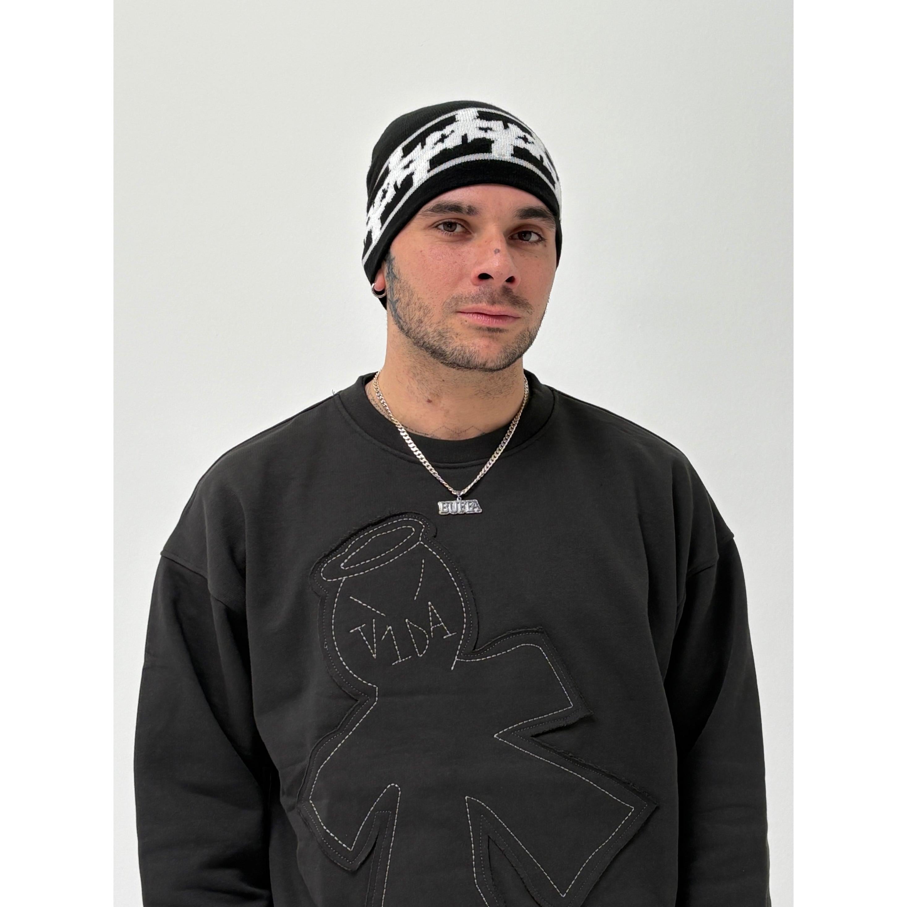 Wearing the V1da Pattern Beanie in black and white low fit, a person pairs it with a black sweatshirt featuring a stitched figure design and a silver necklace, standing against a plain white background.