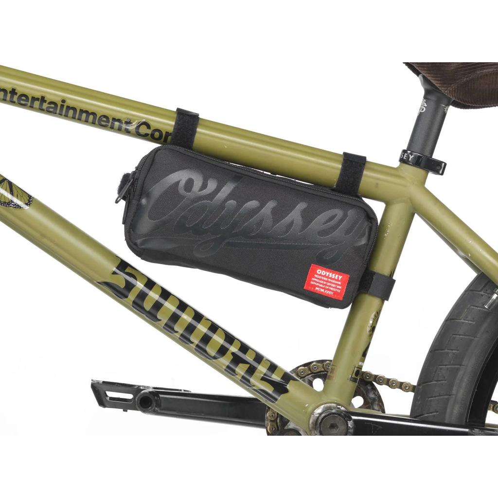 An ODYSSEY Switch Pack V2 Hip Bag in black is secured to the frame of an olive green bike above the pedals with Velcro straps.