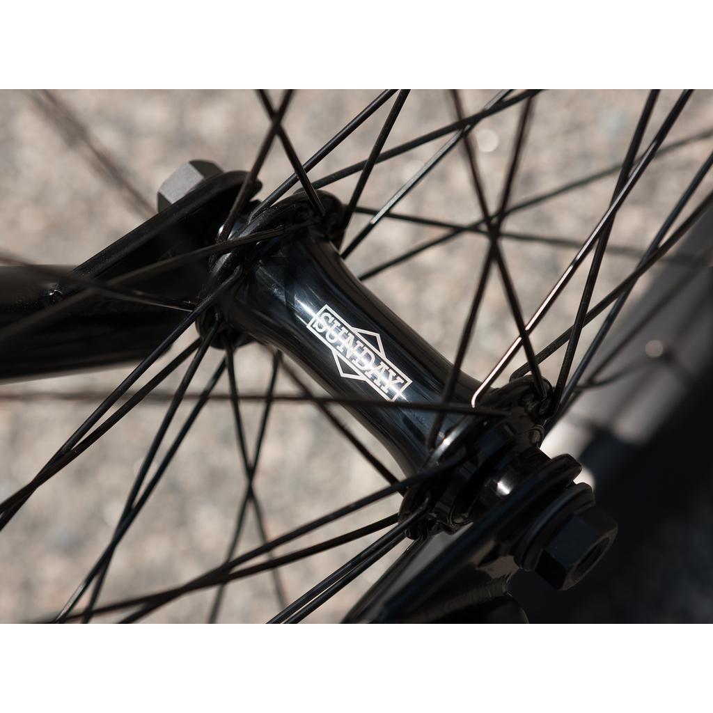 Close-up of a black bicycle hub with the "Shadow" logo, spokes radiating outward, and a blurred gray background—ideal for the Sunday Forecaster 20 Inch Bike.