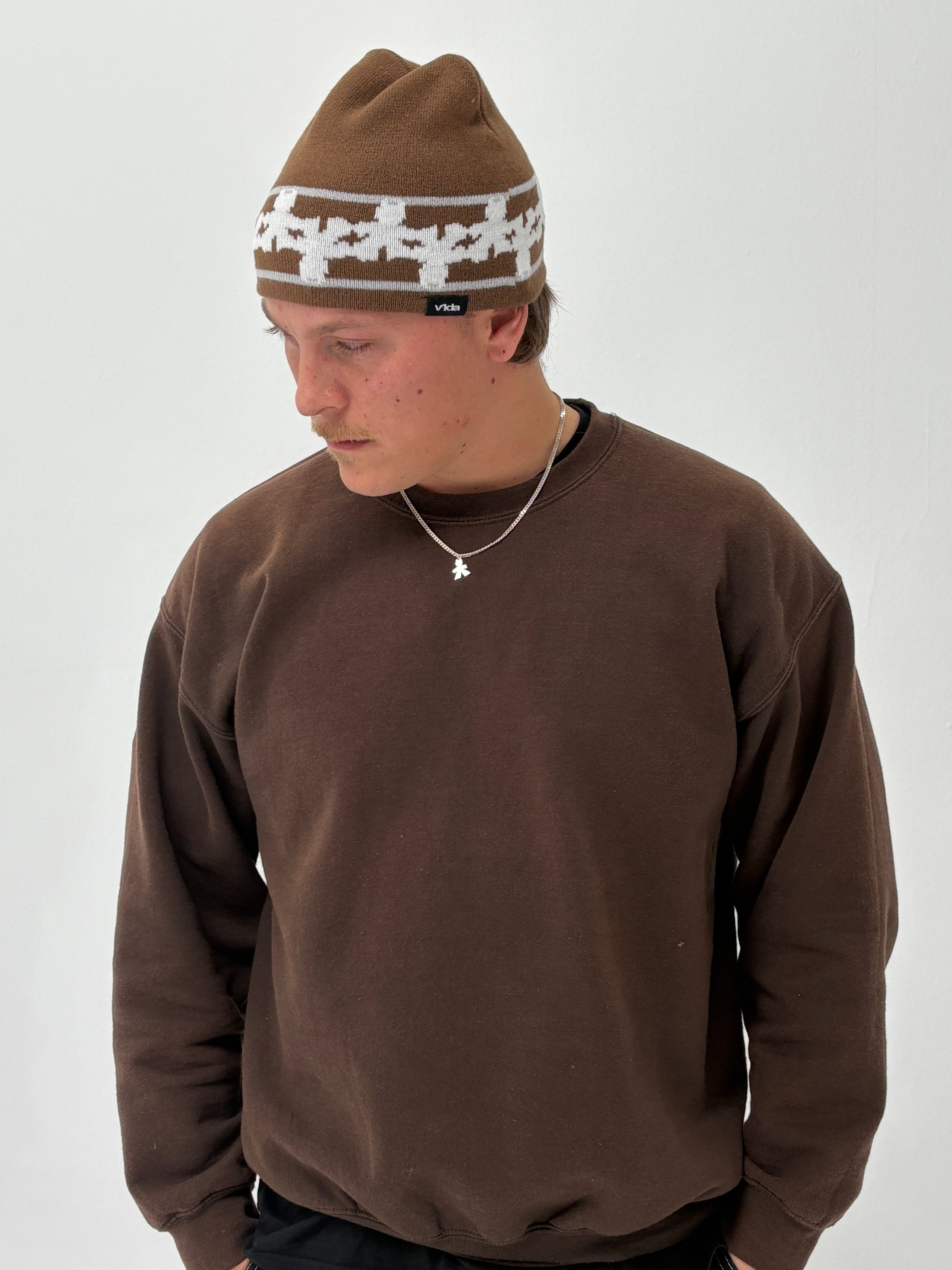 Wearing the V1da Pattern Beanie in brown, a person stands against a plain white background, paired with a brown sweatshirt and small necklace, looking down.