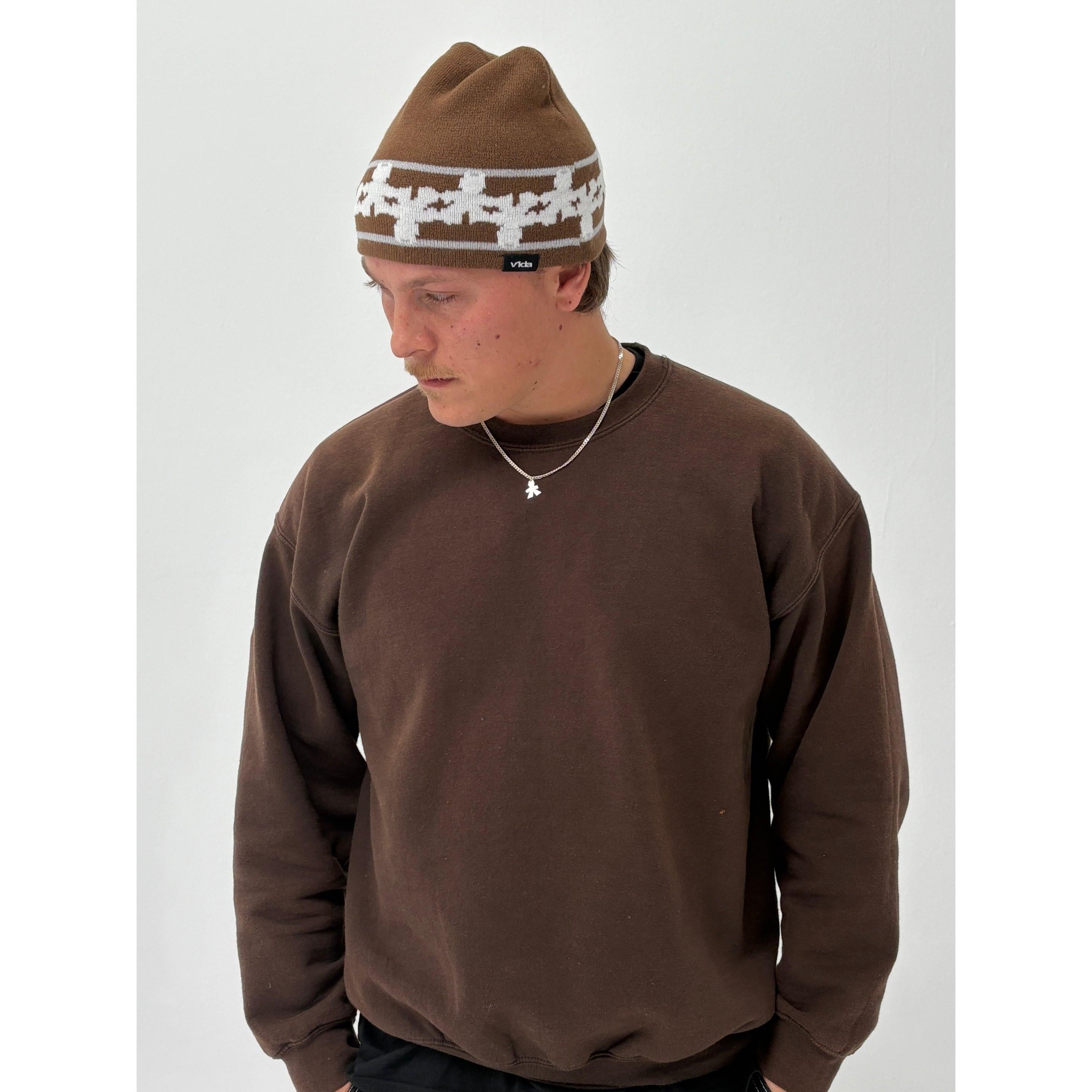 Wearing the V1da Pattern Beanie in brown, a person stands against a plain white background, paired with a brown sweatshirt and small necklace, looking down.