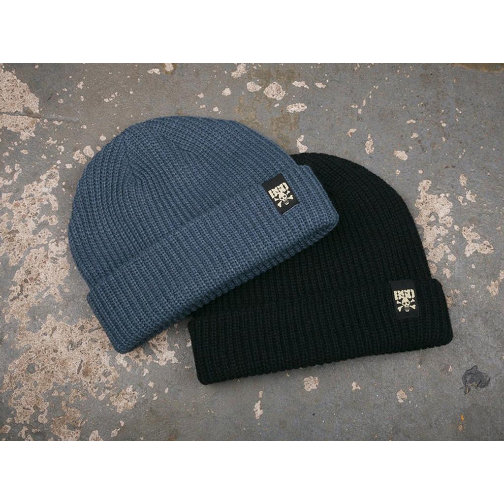 Two BSD More Speed Beanies, one blue and one black, are displayed on a worn concrete surface. Each acrylic knit beanie features a small rectangular logo tag on the cuff.