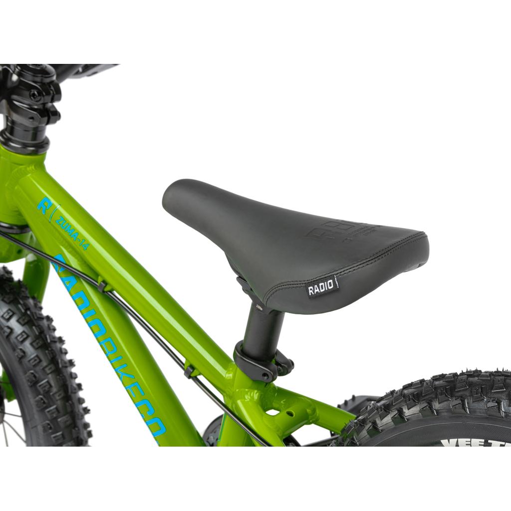 Close-up of a black "RADIO" seat on a green Radio Zuma 14 Inch Bike frame, with part of the rear wheel visible—a lightweight mountain bike designed for kids.
