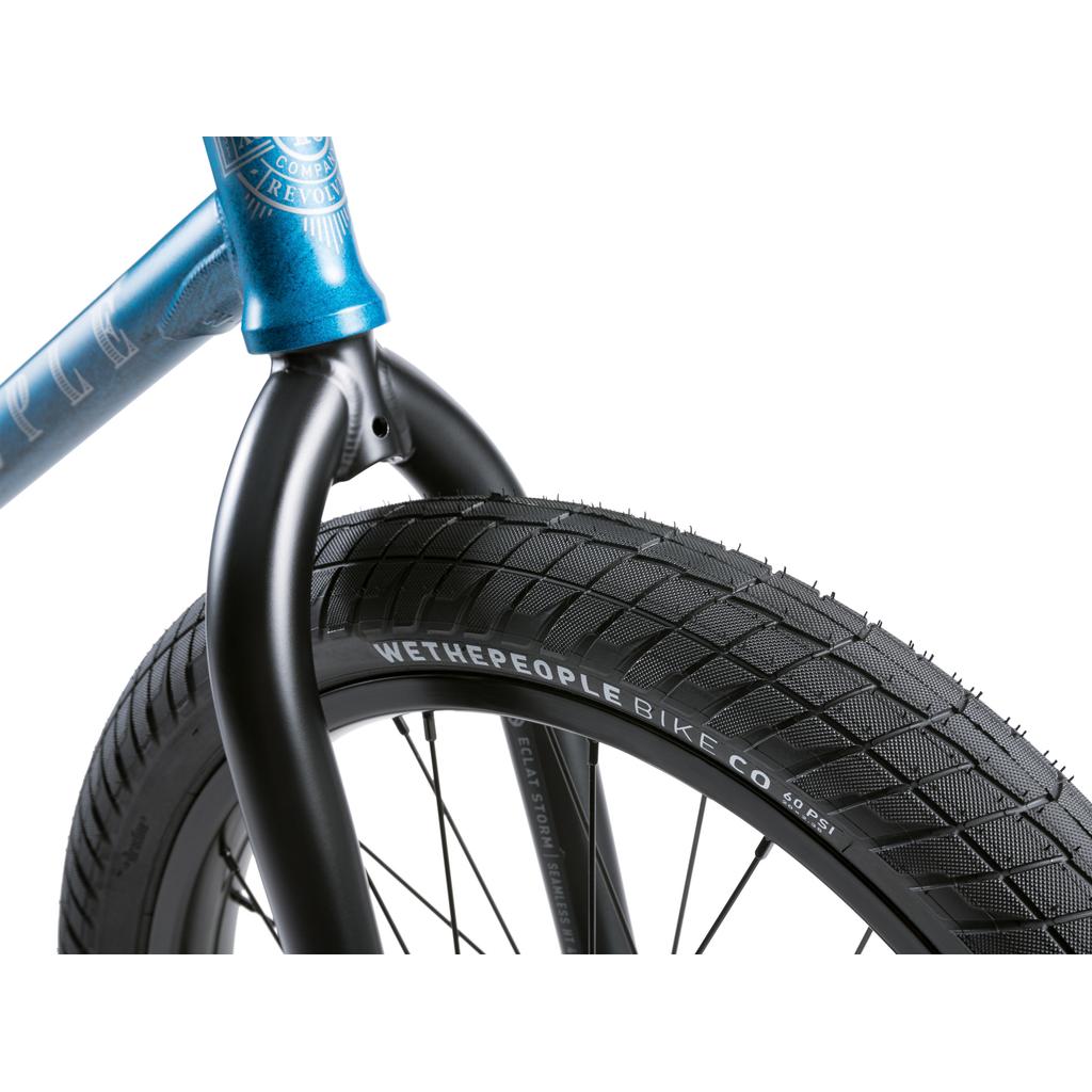 Close-up of the Wethepeople Revolver 20 Inch Bike's front wheel and fork, featuring a thick treaded tire labeled "Wethepeople Bike Co." on a black rim, attached to its blue frame.