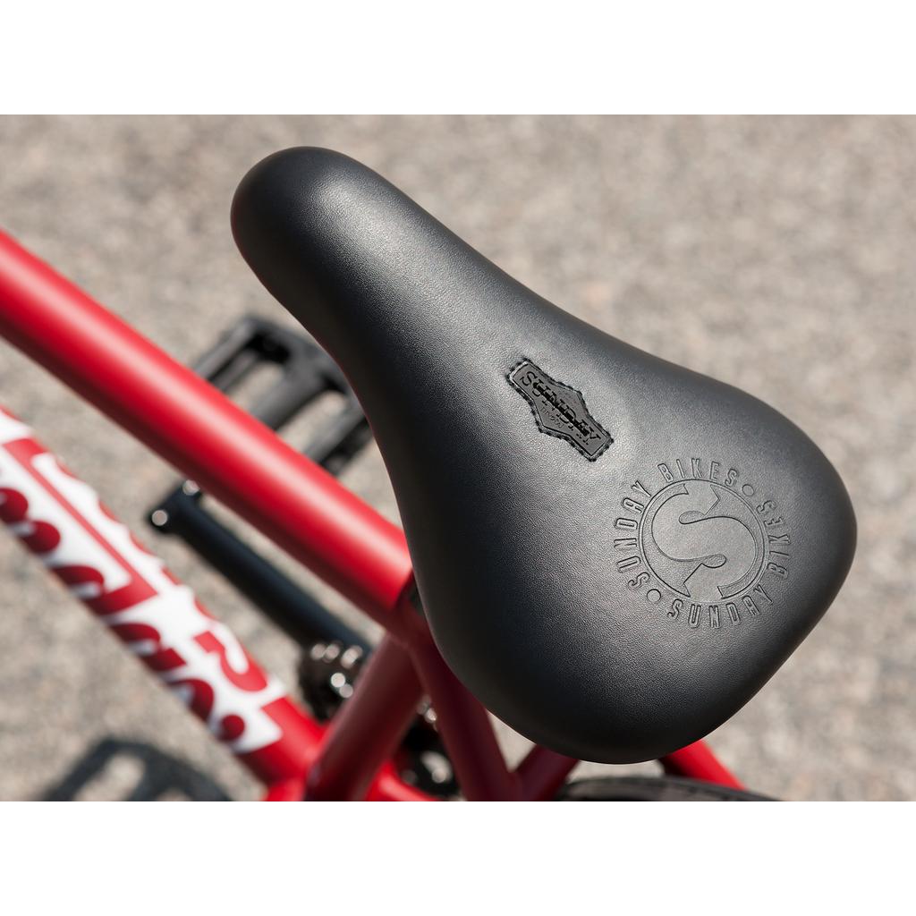 Close-up of a black seat with embossed branding, mounted on a Sunday Forecaster 20 Inch Bike with a red chromoly frame, photographed outdoors on pavement.