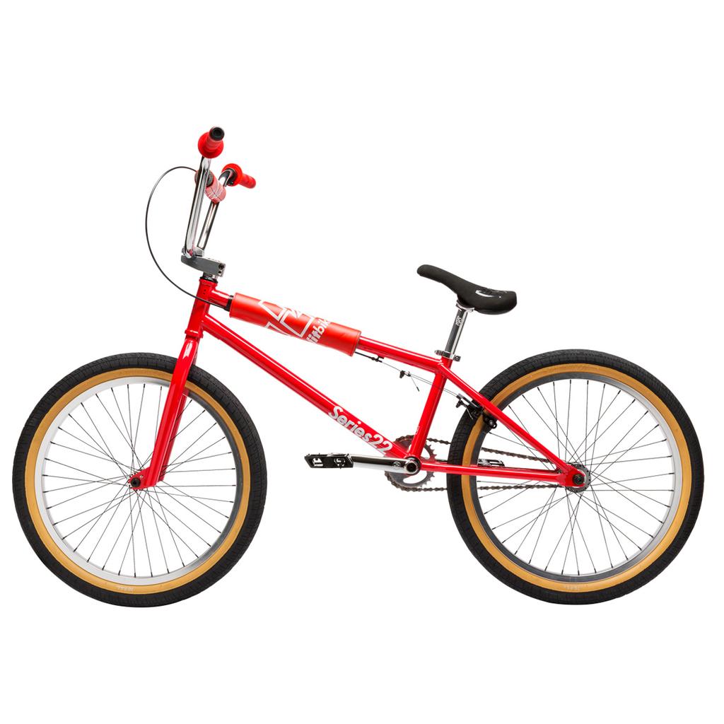 The Fit Bike Co Series 22 Inch Bike features a red frame, black seat, and tan-walled tires, shown from the side against a white background. Its sleek design and bold colors make this BMX bike stand out.