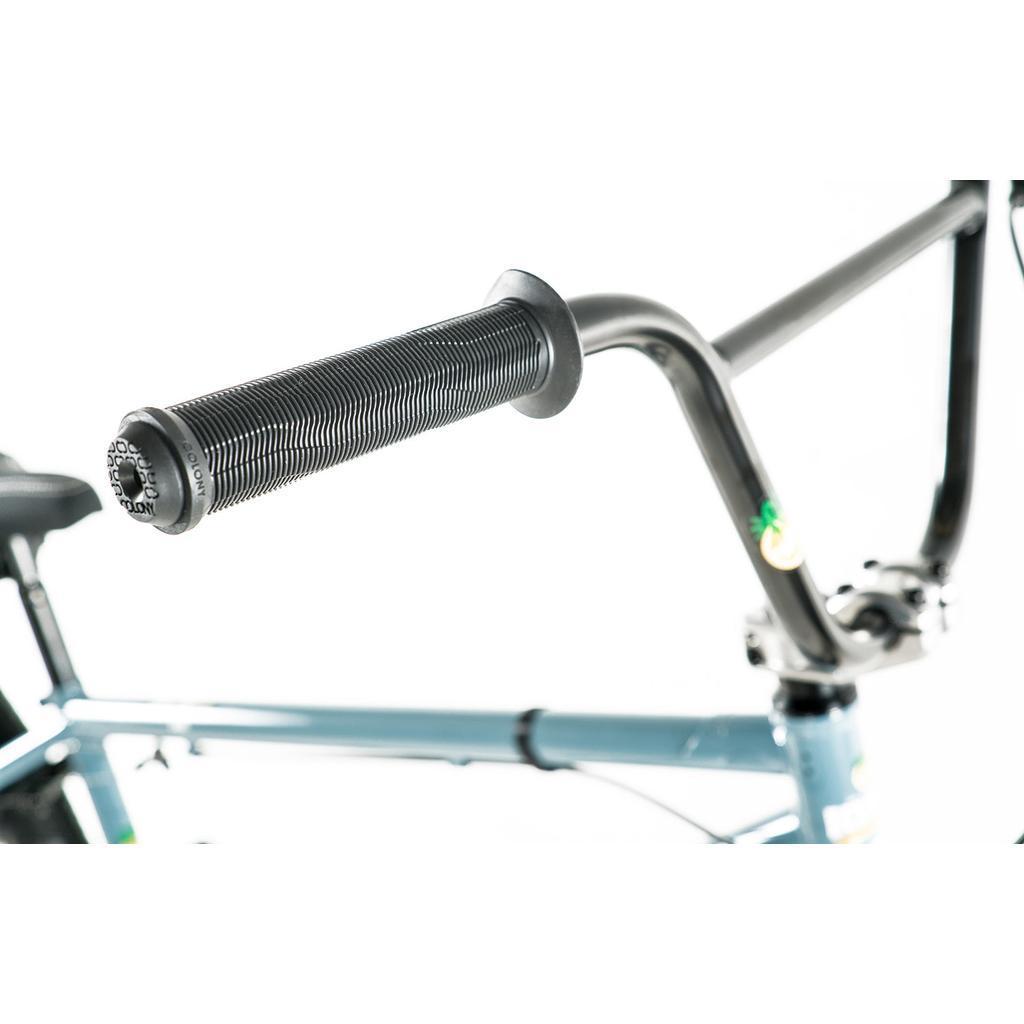 Close-up of a black BMX handlebar grip with the blue Colony Eclipse 26 Inch Bike’s frame, seat, and stem visible in the background on a white surface.