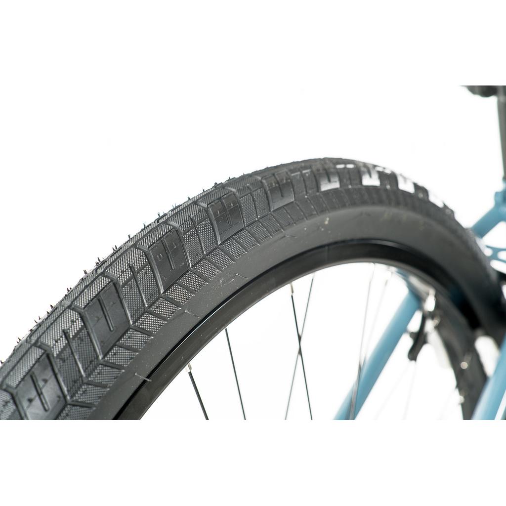 Close-up of a black tire with visible tread, mounted on the blue chromoly frame of a Colony Eclipse 26 Inch Bike against a white background.