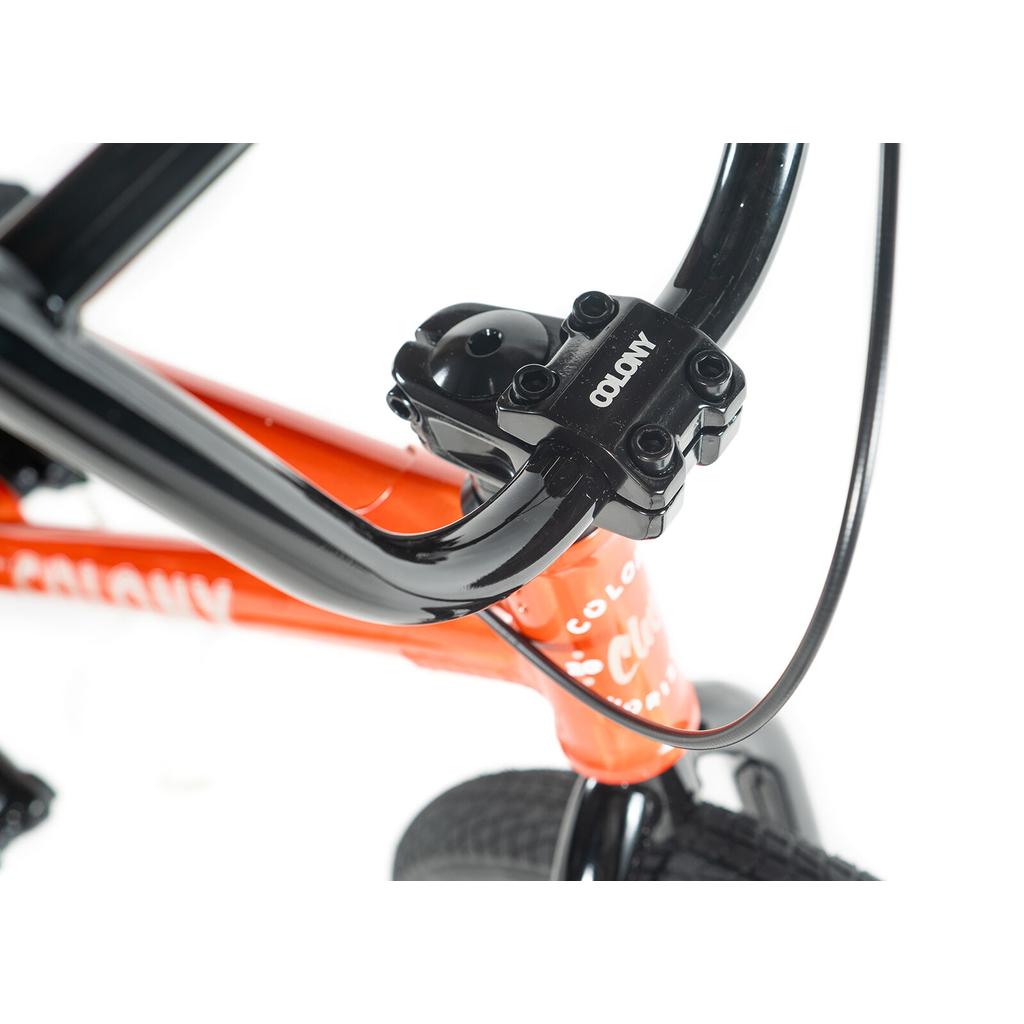 Close-up of the black handlebar and stem with the Colony logo on an orange Colony Horizon 18 Inch Bike, highlighting its lightweight alloy frame, front tire, and brake cable.