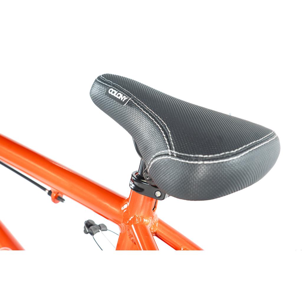 Close-up of a black textured bicycle seat with white stitching on an orange Colony Horizon 18 Inch Bike frame, isolated on a white background.
