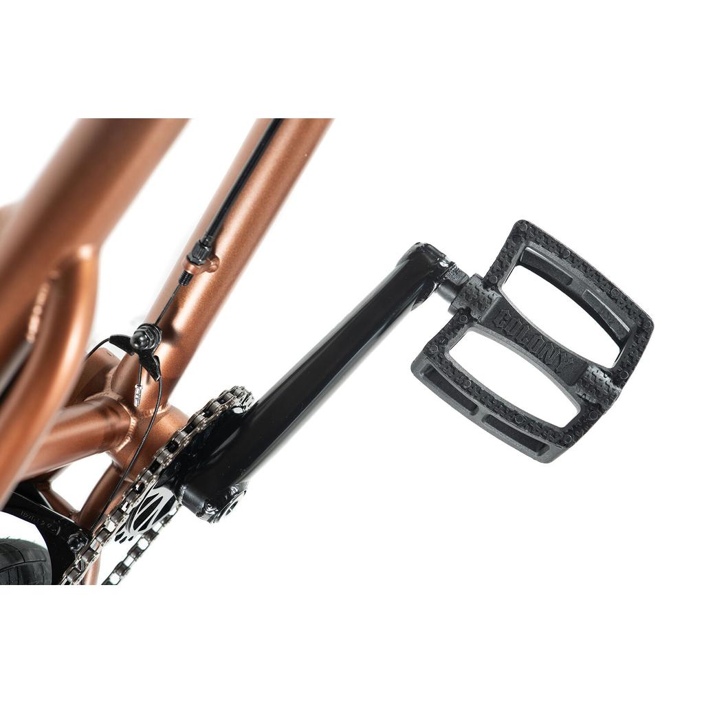Close-up of a black BMX pedal on a crank arm, with the metallic bronze CrMo frame of the Colony Prody Elite 20 Inch Bike in the background.