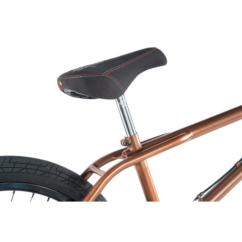 Close-up of a black bicycle seat with red and green stitching, mounted on the metallic brown CrMo frame of the Colony Prody Elite 20 Inch Bike, with part of the rear tire visible.