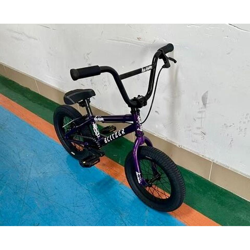 A Division Blitzer 14 Inch Bike in purple, with black handlebars and pedals, sits on a blue and green indoor sports floor beside a white wall.