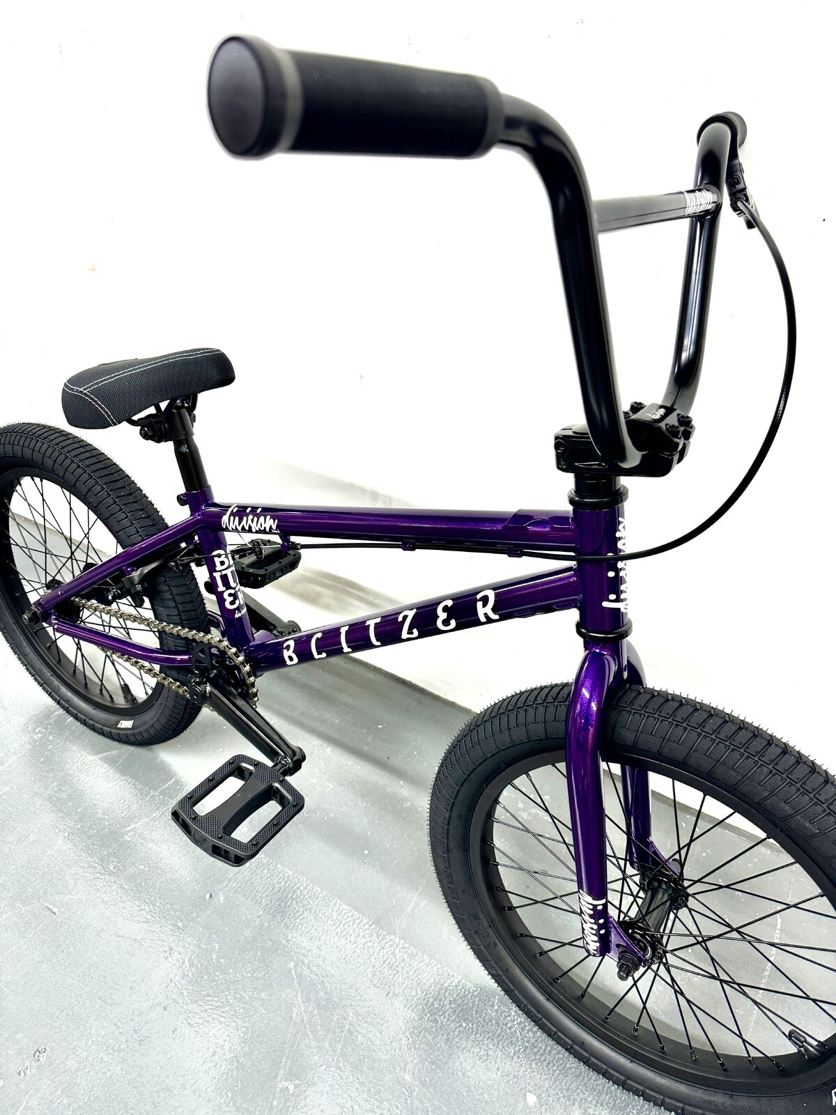 The Division Blitzer 16 Inch Bike, featuring a sturdy Hi-Ten frame with black handlebars, seat, and tires in purple, is pictured on a gray concrete floor against a white wall.