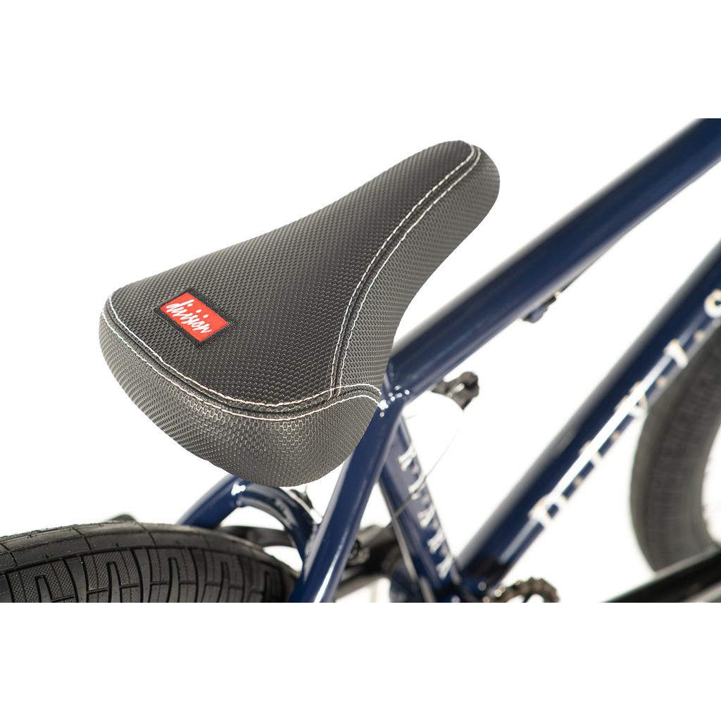 Close-up of a gray textured seat with a red and white logo, mounted on the lightweight Hi-Tensile steel frame of the Division Reark 20 Inch Bike in dark blue, near the rear tire.