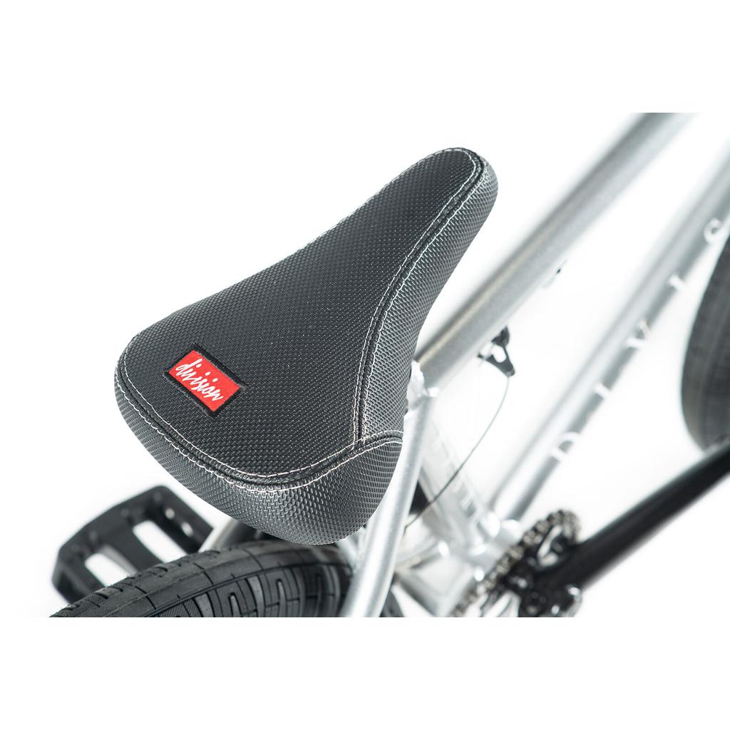 Close-up of a black textured seat with white stitching and a red label, mounted on the lightweight Hi-Tensile steel frame of the Division Reark 20 Inch Bike, with the rear tire also visible.