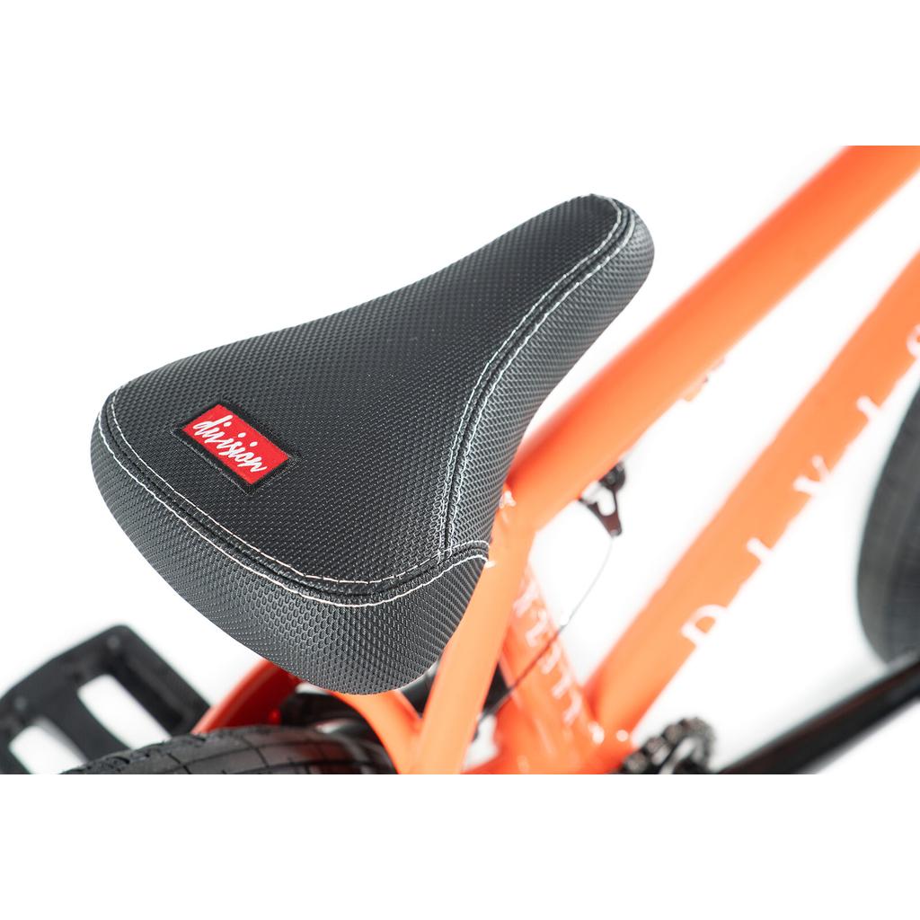 Close-up of a black textured bike seat with white stitching, mounted on an orange Division Reark 20 Inch Bike frame made of lightweight Hi-Tensile steel; a red tag labeled "Mission" is sewn onto the seat.