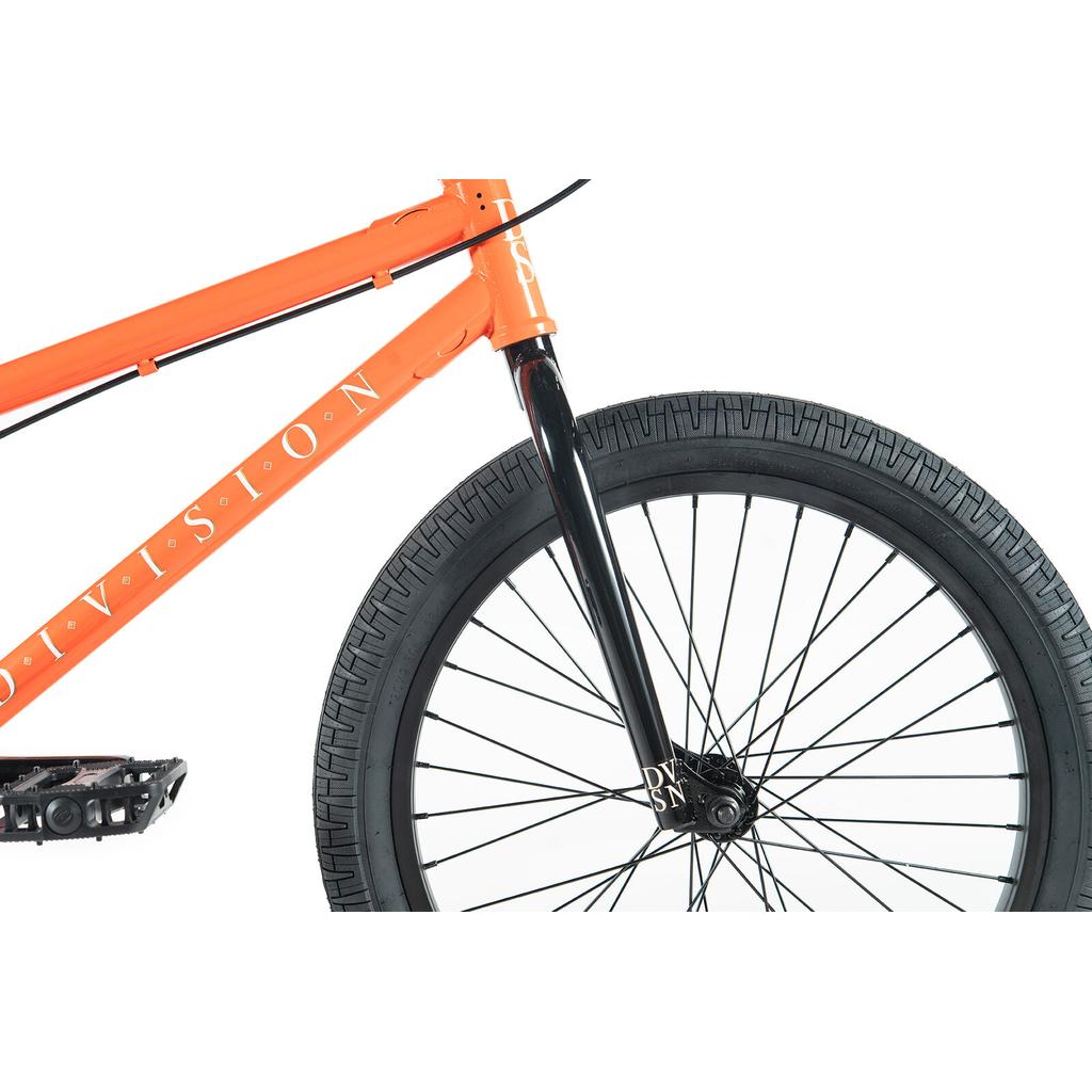 Close-up of the front section of a Division Reark 20 Inch Bike in orange, highlighting the frame, black front fork, wheel, tire, and part of the pedal against a white background.