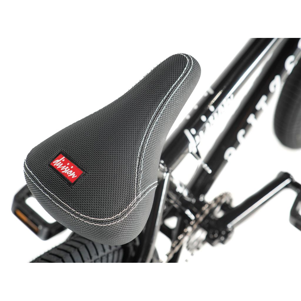 Close-up of a black seat with white stitching and a red "division" tag, mounted on the Division Blitzer 16 Inch Bike's black frame near the rear tire.
