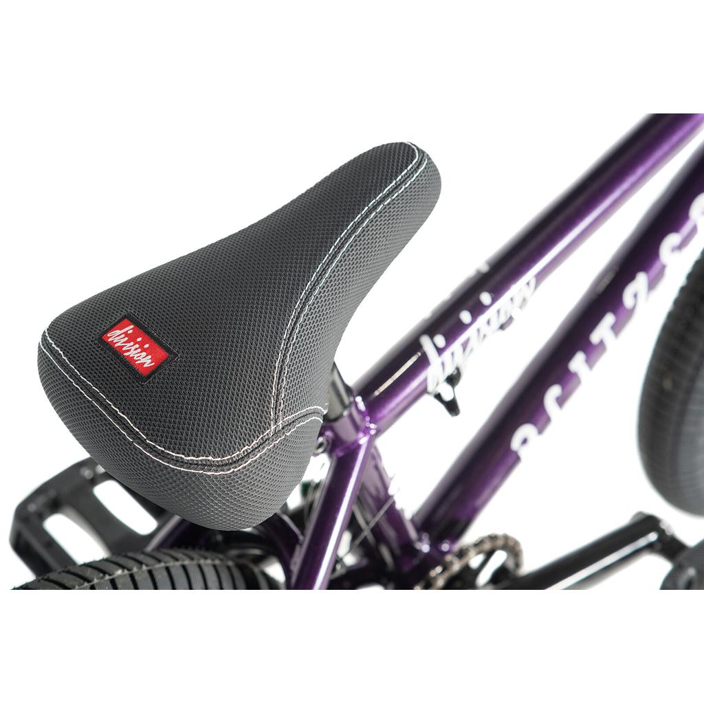 Close-up of a black textured seat with white stitching and a red label on the Division Blitzer 16 Inch Bike, featuring a purple frame and 16 inch wheels.
