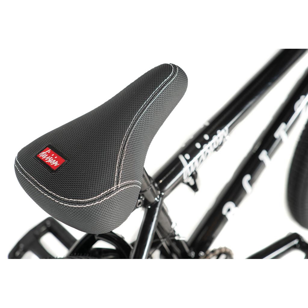 Close-up of a black bicycle seat with white stitching and a red label on the Division Blitzer 18 Inch Bike—ideal for beginner BMX riders or kids—shown against a white background.